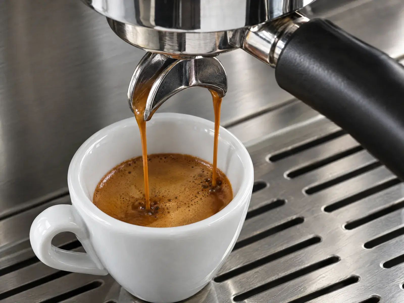 Two streams of espresso pouring into a white ceramic cup on a steel drip tray